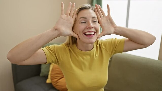 Cheerful young blonde woman at home, capturing hearts with her sparkling eyes, happily playing peek a boo showing face, wearing casual t-shirt and sharing excited, joyful moments indoors.
