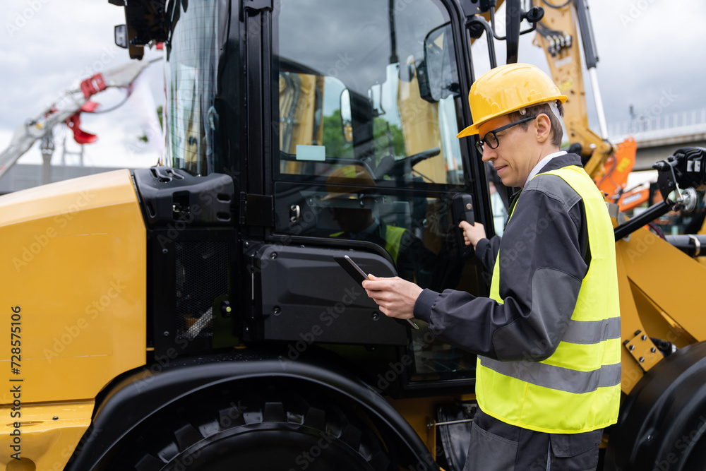 Poster engineer in a helmet with a digital tablet stands next to construction excavators..