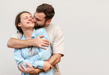 Happy young woman in the arms of her husband in front of light grey background win studio. Man hugs woman.