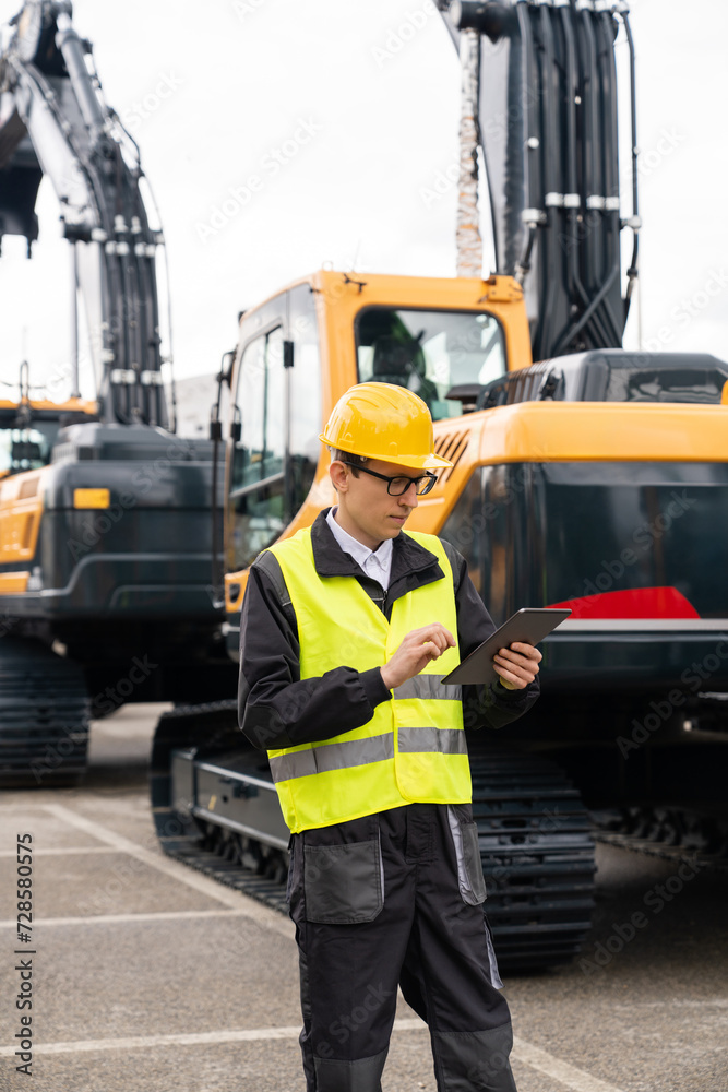 Poster engineer in a helmet with a digital tablet stands next to construction excavators..