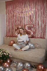 Happy young woman blowing birthday candle at home. birthday girl with a celebratory cake for twenty-five years. pink photo zone with balloons for birthday. The happy woman makes a wish