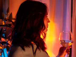 A sexy young woman in a dress holds a glass of white wine. Home portrait, neon light.
