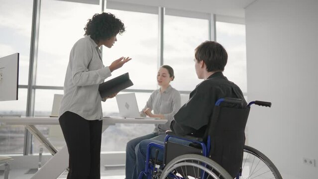Paralyzed man works with colleagues. Guy in wheelchair has spine trauma discussing working project in office on meeting with multiethnic team. Teamwork brainstorming, special possibilities needs.