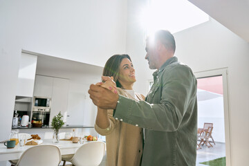 Happy affectionate romantic middle aged mature couple dancing at home. Smiling senior older man and woman in love standing in house kitchen enjoying dance lit with sunlight. Authentic photo.
