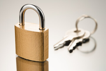 Closed metal padlock in golden color and two keys on a reflective surface. Front view from low...