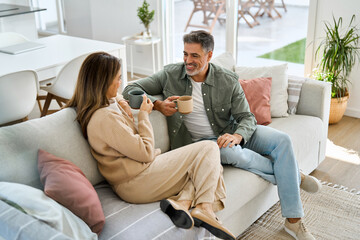 Happy middle aged mature couple talking drinking coffee relaxing on couch at home. Relaxed older...