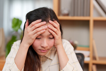 Distress and fatigue shadow face of Asian office worker as grips head, overwhelmed by work. Asian businesswoman manifests stress, hands on temples, feeling weight of work pressures in modern office