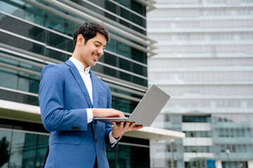 Fototapeta premium A smiling Hispanic businessman stands with an open laptop in front of a modern office, embodying the concept of accessible technology and business efficiency in today's fast-paced world.