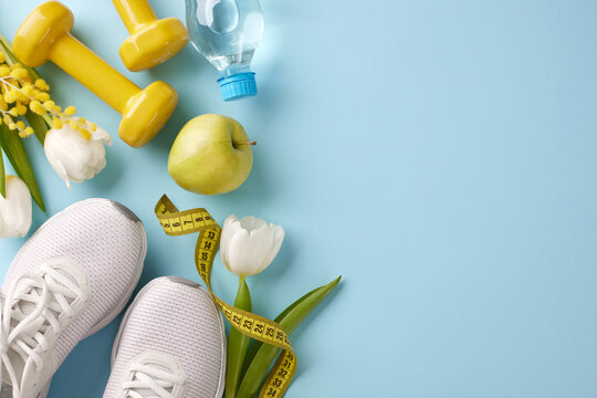 Seasonal fitness renewal: transform your routine this spring. Top view photo of white sneakers, yellow dumbbells, measure tape, flowers, water bottle on pastel blue background with advert area