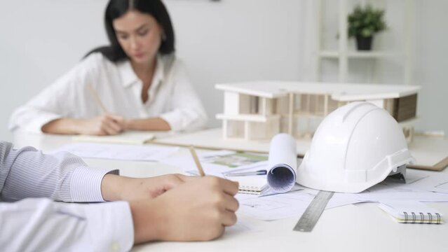 Professional architect engineer drawing blueprint and working together with young beautiful caucasian coworker on meeting table with safety helmet, house model, blueprint scatter around. Immaculate.