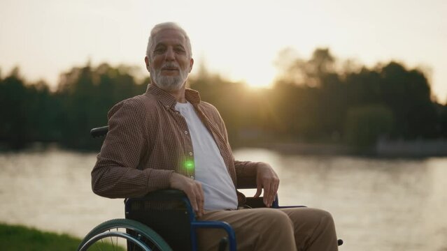 Senior man in wheelchair enjoys freedom. Male with special need look at the camera smiling breathing fresh air walking in city park near nature. Living with physical disability in old age concept.