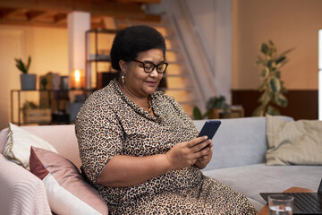 Portrait of modern Black senior woman wearing cheetah print dress using smartphone sitting on sofa in home interior copy space - obrazy, fototapety, plakaty