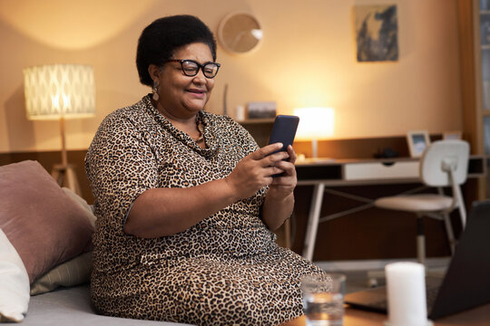 Side view portrait of smiling senior African American woman holding phone and wearing glasses at home while using online messaging apps copy space