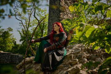 Mittelalterliche Frau mit roten Haaren als Kriegerin mit Kriegsbemalung mit Pfeil und Bogen im Sommer vor einer alten Burgruine, mit Korsett aus Leder und Umhang 