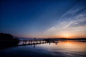Silhouette of a person on a pier against the twilight sky reflected on a calm lake's surface