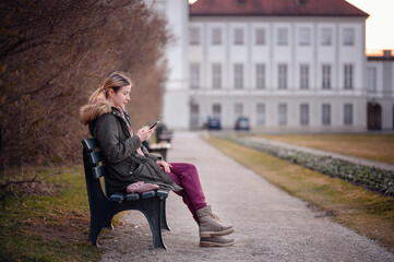 A young woman sits on a park bench, lost in thought with a smartphone in her hand