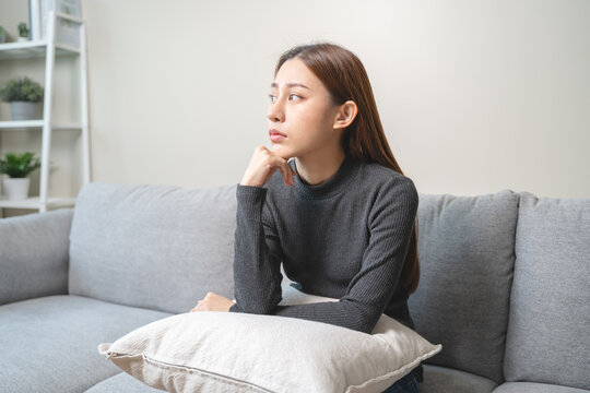 Unhappy anxiety young Asian woman covering her face with knee on the sofa in the living room at home.