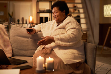 Side view portrait of senior African American woman enjoying video chat talk with friend and drinking wine in cozy home setting copy space