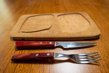 Wooden board with cutlery (knife and fork) both on wooden table.