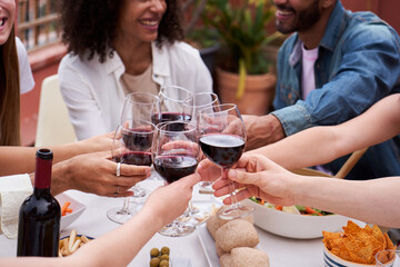 Close up view of a group of unrecognizable people toasting with red wine outdoors, having fun enjoying the day together with food and drinks. Multiracial friends gathering to celebrate.