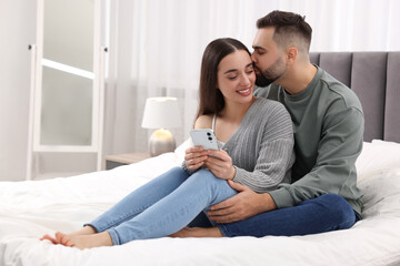 Happy young couple with smartphone in bedroom. Space for text