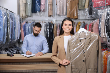 Dry-cleaning service. Happy woman holding hanger with coat indoors. Worker taking notes at workplace