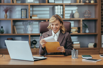 Happy senior business woman in brown suit is sitting in the office at the desk and opening the...