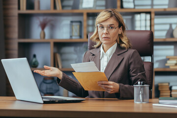 Portrait of a worried senior businesswoman in a business suit sitting in the office at a desk, holding a letter in an envelope and looking disappointed at the camera.
