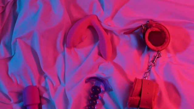 set of woman sex toys for masturbation and orgasm on sheet on bed. Female vibrator, vacuum stimulator, butt plug and BDSM handcuffs in neon lighting