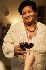 Vertical portrait of smiling African American mature woman clinking wine glasses with unrecognizable daughter visiting mother