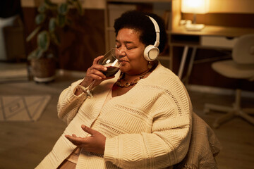 Portrait of senior African American woman drinking wine at home and listening to music with headphones, copy space