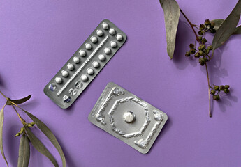 Two different packs of contraceptive pills on colored paper background with eucalyptus leaves,...
