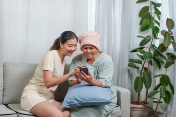 young Asian woman guides elderly, ailing mother through a smartphone, sharing a connection. tender scene unfolds as young daughter helps sick mother operate phone, bridging technology gap with care.