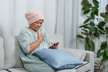 elder woman in pink knit cap feels discomfort, clutching chest while holding phone, alone at home. moment of concern as senior woman experiences pain, expression worried, in a quiet living space