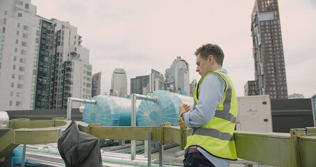 Male engineer in hi-vis vest analyzing equipment on city building rooftop