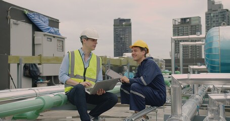 Two engineers in safety helmets and vests are working with a laptop on a rooftop surrounded by HVAC...