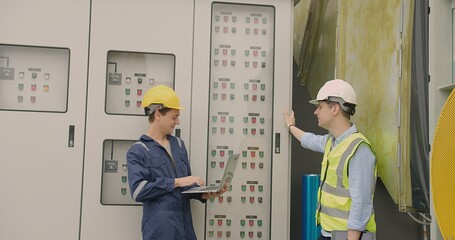 Two engineers in hard hats engage with complex control panel machinery, laptop in hand, sharing...