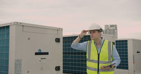 Serious male engineer with a clipboard and hard hat inspecting rooftop HVAC units against a dusky sky