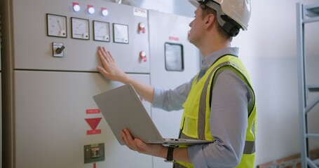 An engineer in a high-visibility vest and hard hat inspects systems on his laptop at an industrial control panel - obrazy, fototapety, plakaty