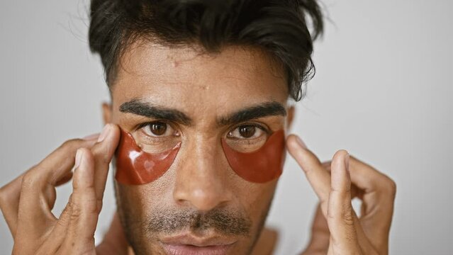 Close-up of a young hispanic man applying skincare patches under eyes against an isolated white background
