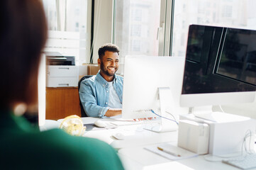 Joyful Indian male employee in casual denim shirt enjoys a light moment at his workstation, showcasing the casual yet professional atmosphere of a modern open space office. Friendly office atmosphere