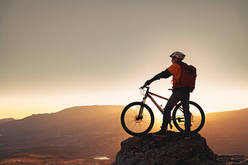 Biker male silhouette is standing with mtb bike at mountain top against sunset sky