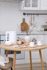 Home spring easter interior in kitchen. Beige chair with white cover, white electric kettle, hot teapot, vase with spring branches candle, cups of tea on round wooden table in cosy white kitchen.