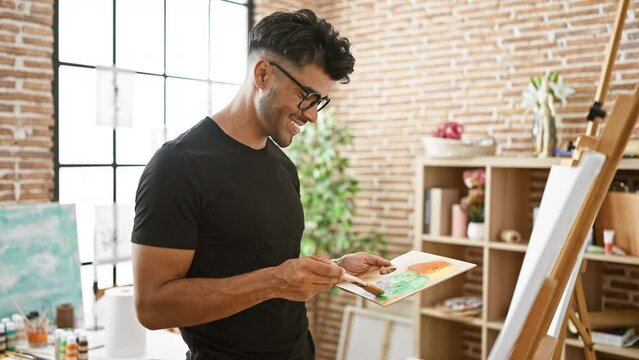 A cheerful young hispanic man painting in a bright art studio, wearing a black shirt and glasses.