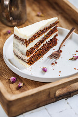 A piece of carrot cake served on rustic plate placed on bright sunny scene. Layered cake with cream cheese frosting and fluffy biscuit. Served with tea. 