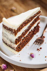 A piece of carrot cake served on rustic plate placed on bright sunny scene. Layered cake with cream cheese frosting and fluffy biscuit. Served with tea. 