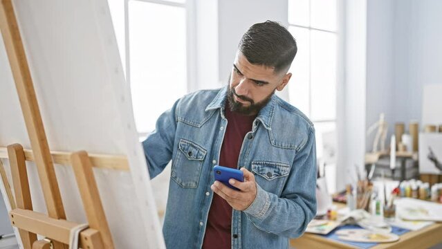 Focused man using smartphone in creative studio with easel and paintings