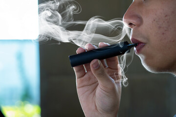 Asian preteen holding and smoking e-cigarette in dark room to escape the view of parents because it...