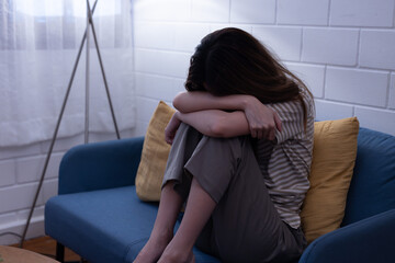 Obraz premium Depressed asian woman covering face with knee sitting on couch