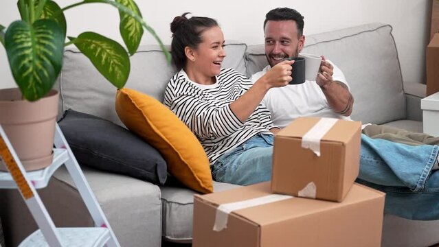 In new home, a young couple on the sofa toasts with coffee cups amid unpacked boxes, savoring the thrill of unboxing and embracing their exciting journey into a new chapter